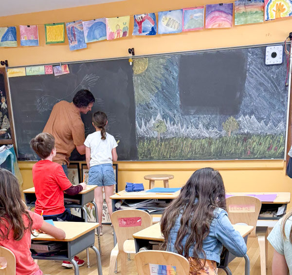 elementary classroom - students work at desks and the teacher is writing on the blackboad