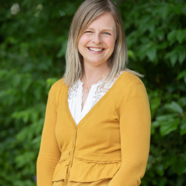 headshot of Anne Coyner at CWS - blonde woman wearing a yellow cardigan posing in front of leafy green trees