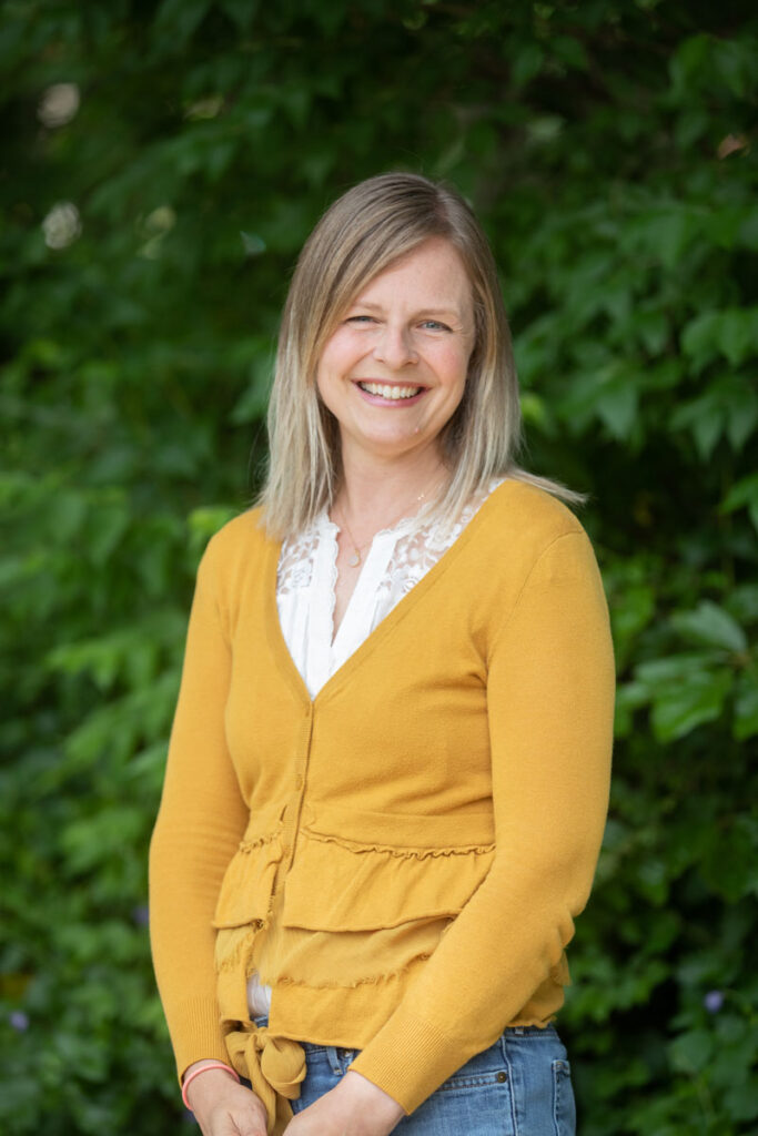 headshot of Anne Coyner at CWS - blonde woman wearing a yellow cardigan posing in front of leafy green trees